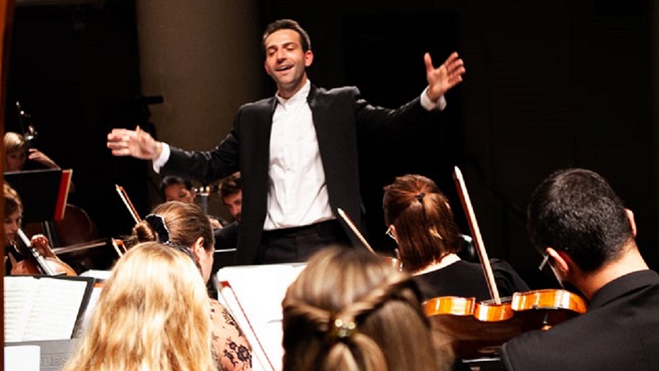 A male alumna, with dark hair, wearing a suit, conducting an orchestra. 